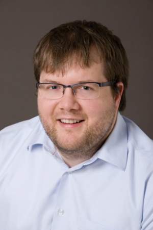 Portrait de Philippe Beauchemin souriant portant une chemise bleu pâle et des lunettes, devant un fond uni gris.