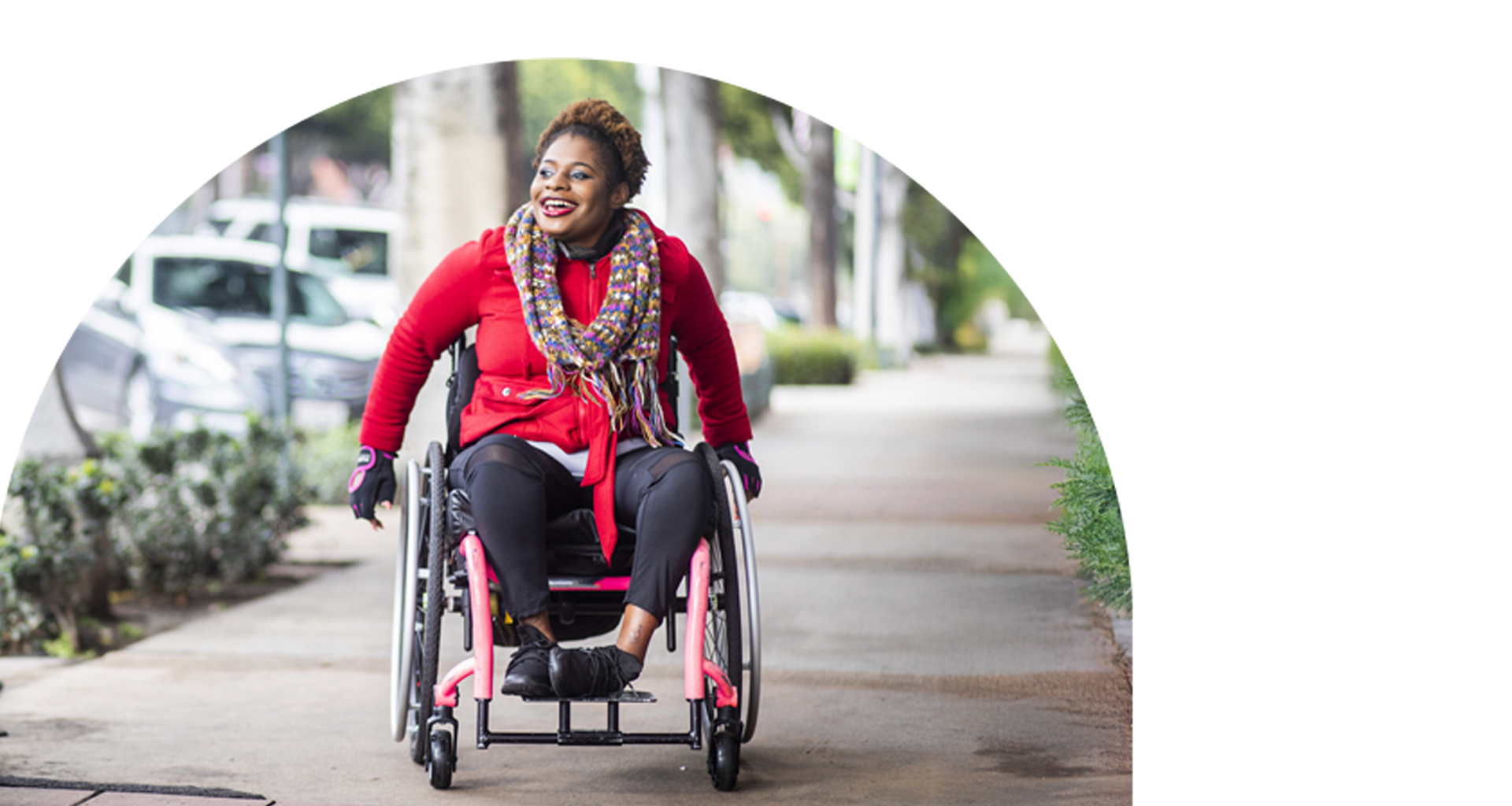 A smiling woman in a wheelchair rolls along a city sidewalk.