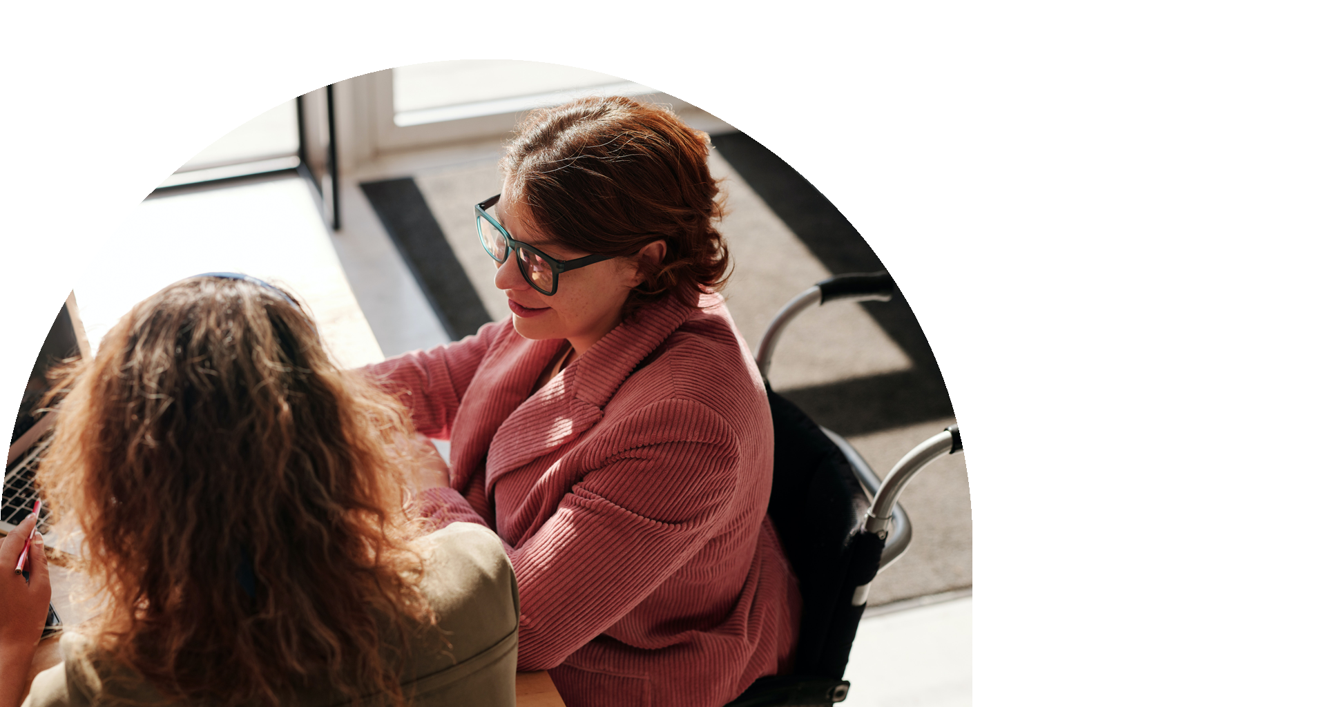 An overhead view of woman with short, reddish hair and glasses in a wheelchair. She is showing another woman her laptop screen.