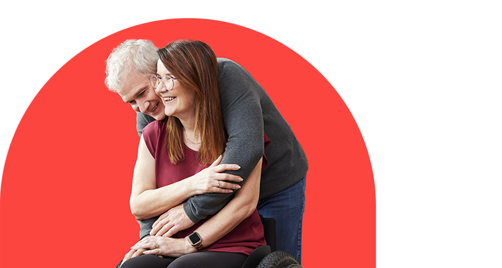 A woman with long brown hair and glasses is wearing a red shirt and sitting in a wheelchair. She is being embraced by a man with short white hair wearing a grey sweater.