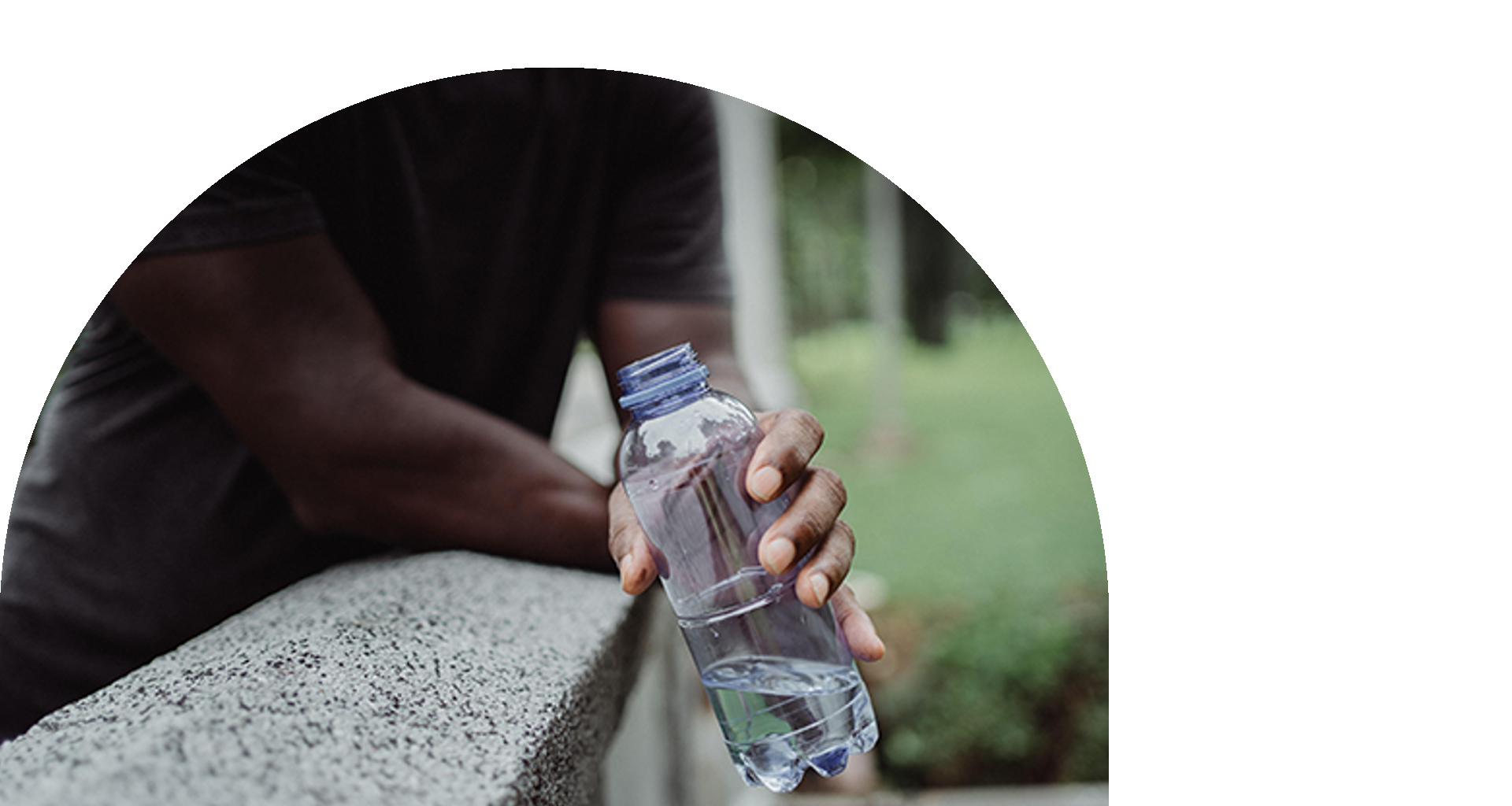 A person in a dark shirt leaning on a concrete surface, holding a clear plastic water bottle with the cap off. 