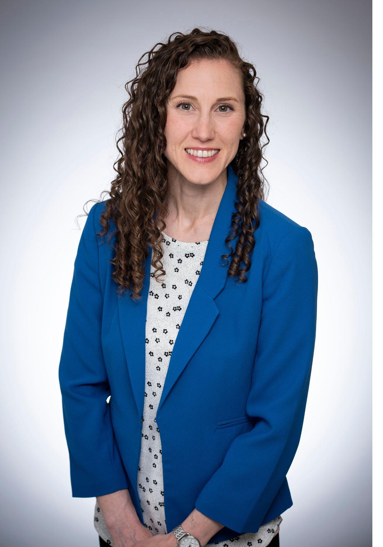 Laura pose en souriant, et elle porte une veste bleu roi par-dessus un chemisier blanc à pois bleus. Elle a des cheveux bruns frisés et des yeux verts.  