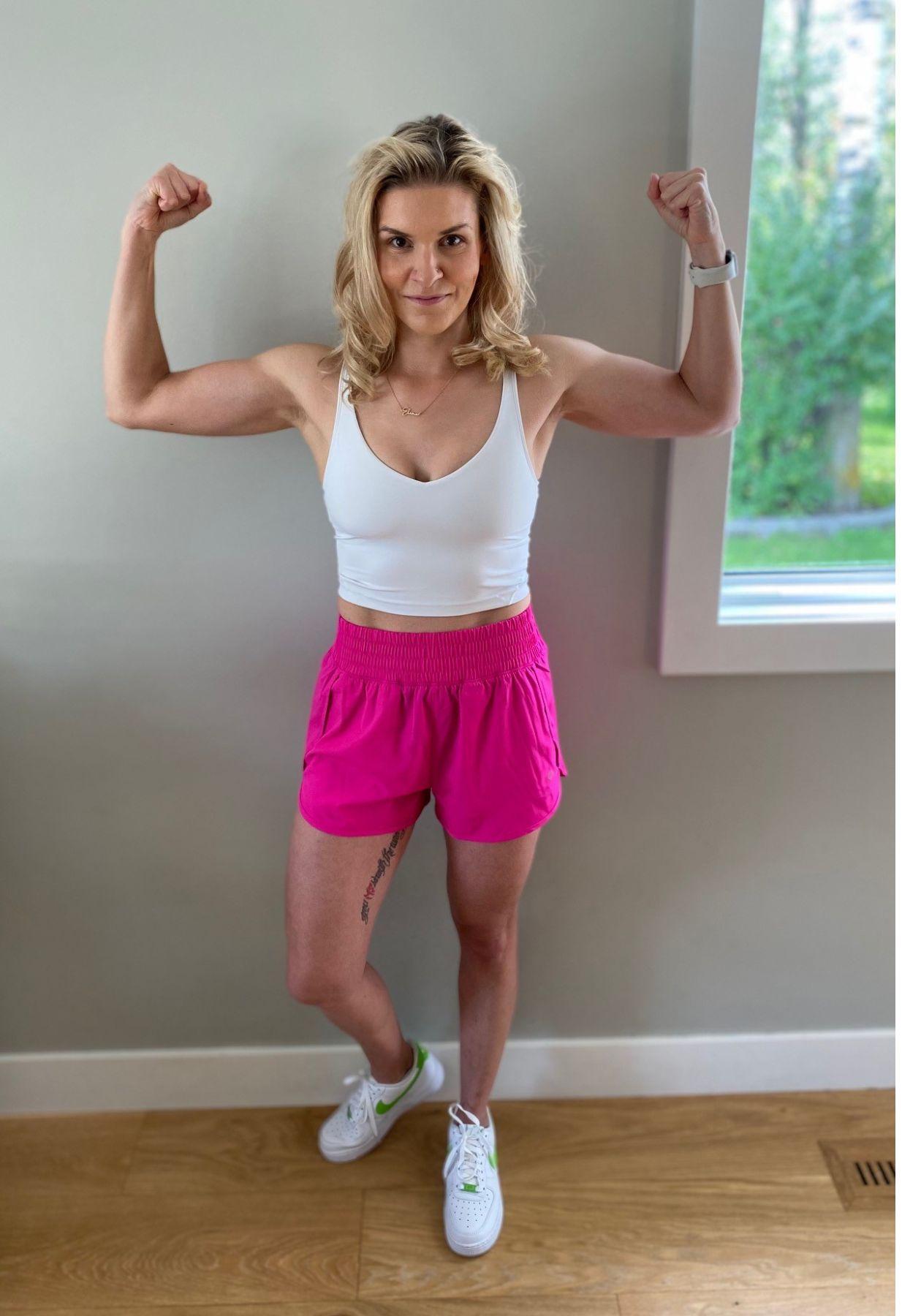 Une femme nommée Patrycia montre les muscles de ses bras. Elle porte un débardeur blanc et une culotte de sport rose vif, et elle est chaussée d’espadrilles blanches.  