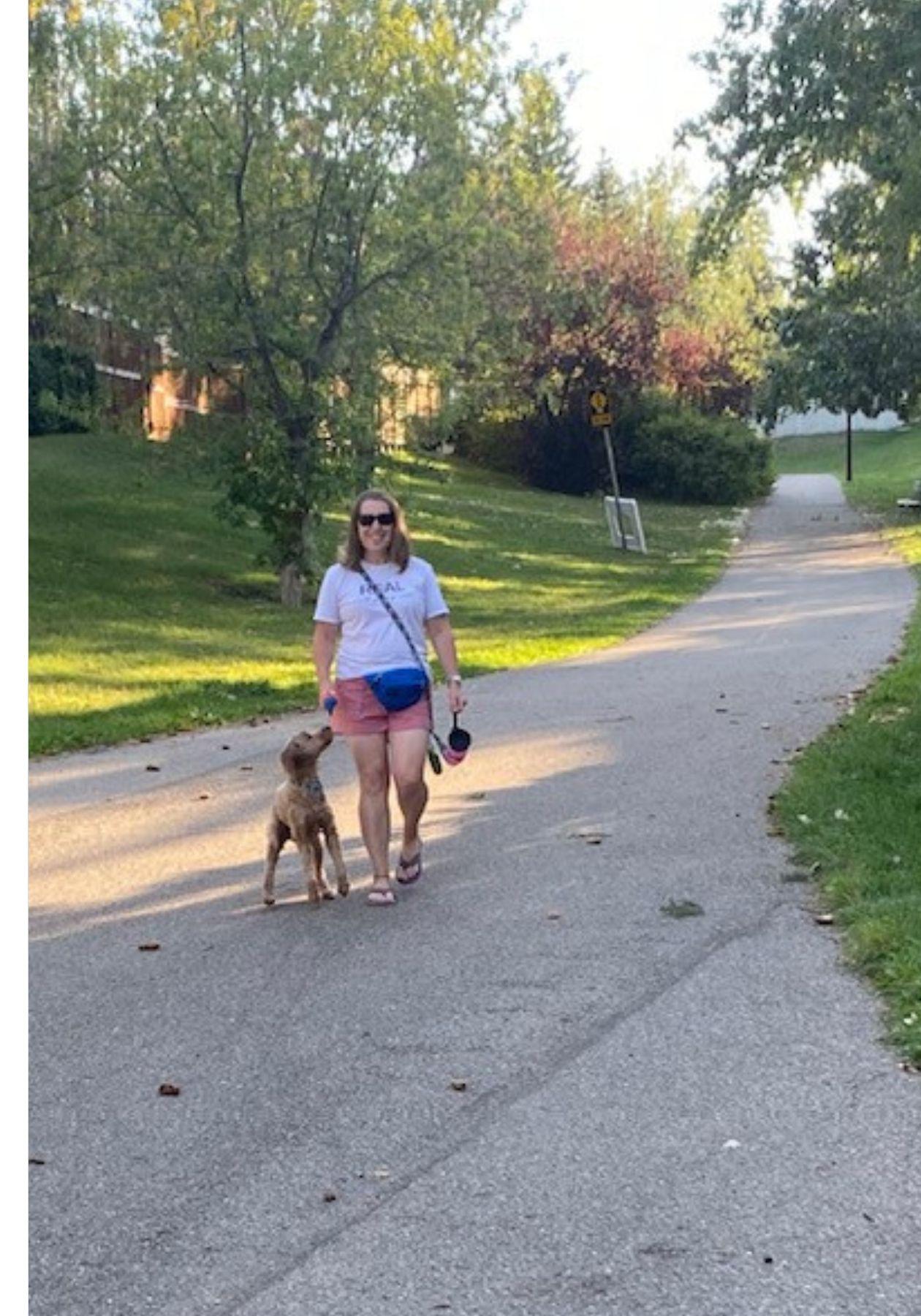 Shauna est vêtue d’un t-shirt blanc et d’un short rouge, et elle porte une sacoche de ceinture bleue et des sandales de plage violettes. Elle promène son chien, qu’elle tient en laisse, dans un quartier résidentiel. 