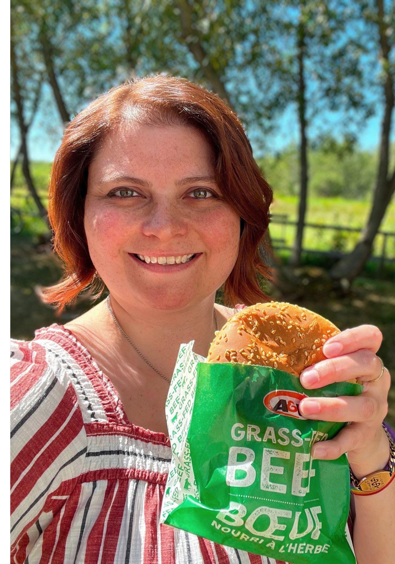 Caitlin sourit en tenant un Teen Burger™. Elle est à l’extérieur. En arrière-plan apparaissent des arbres.