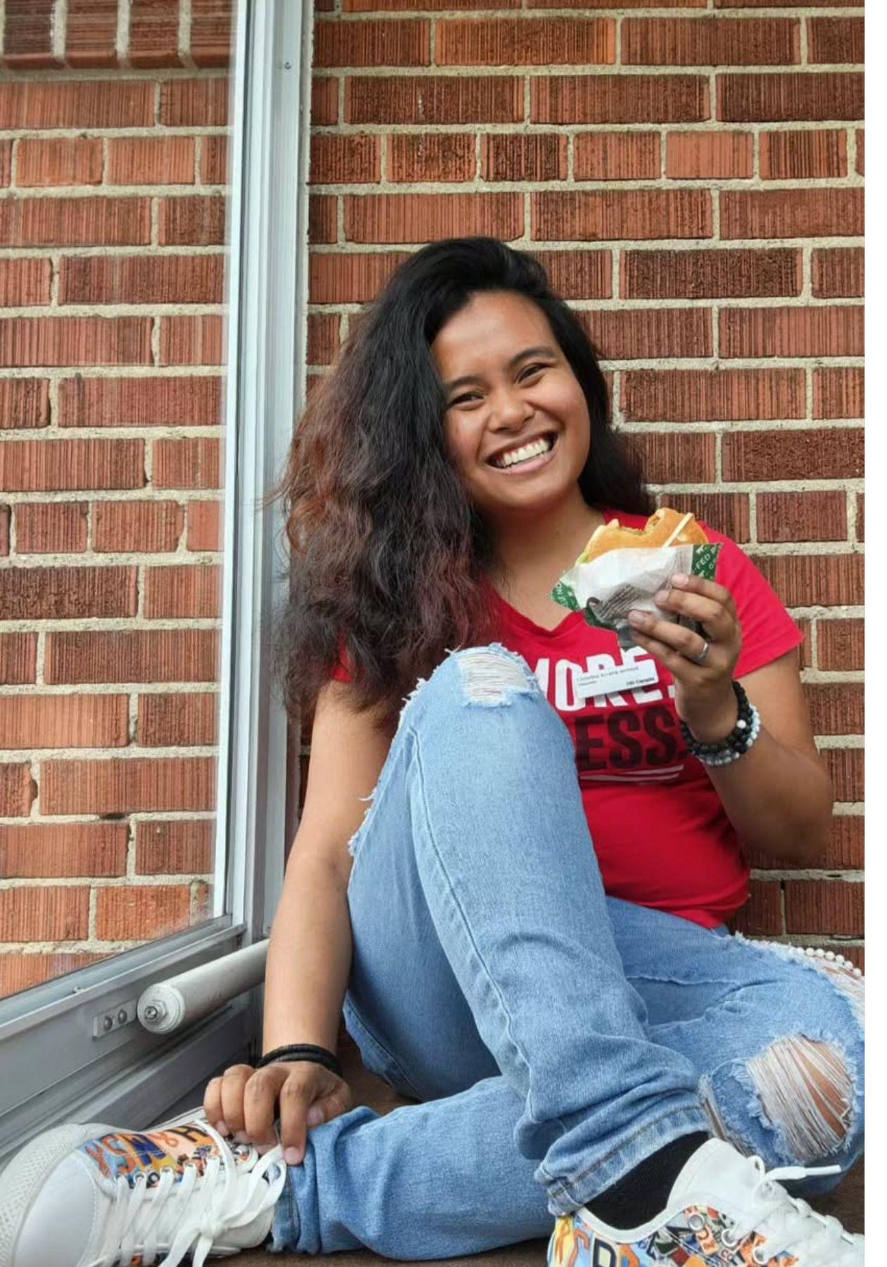 Christina est assise par terre, elle sourit et tient un Teen Burger™.