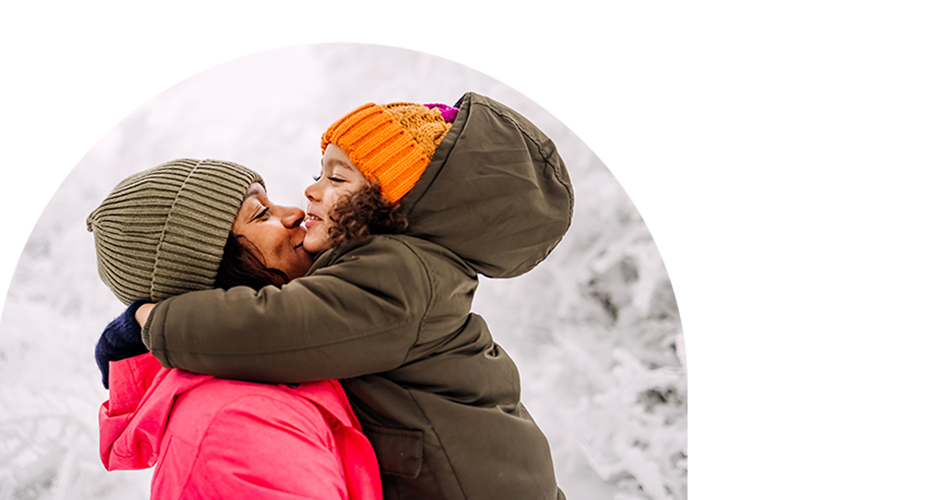 A woman and her young daughter embrace. They are outside in the snow with winter coats and hats on.