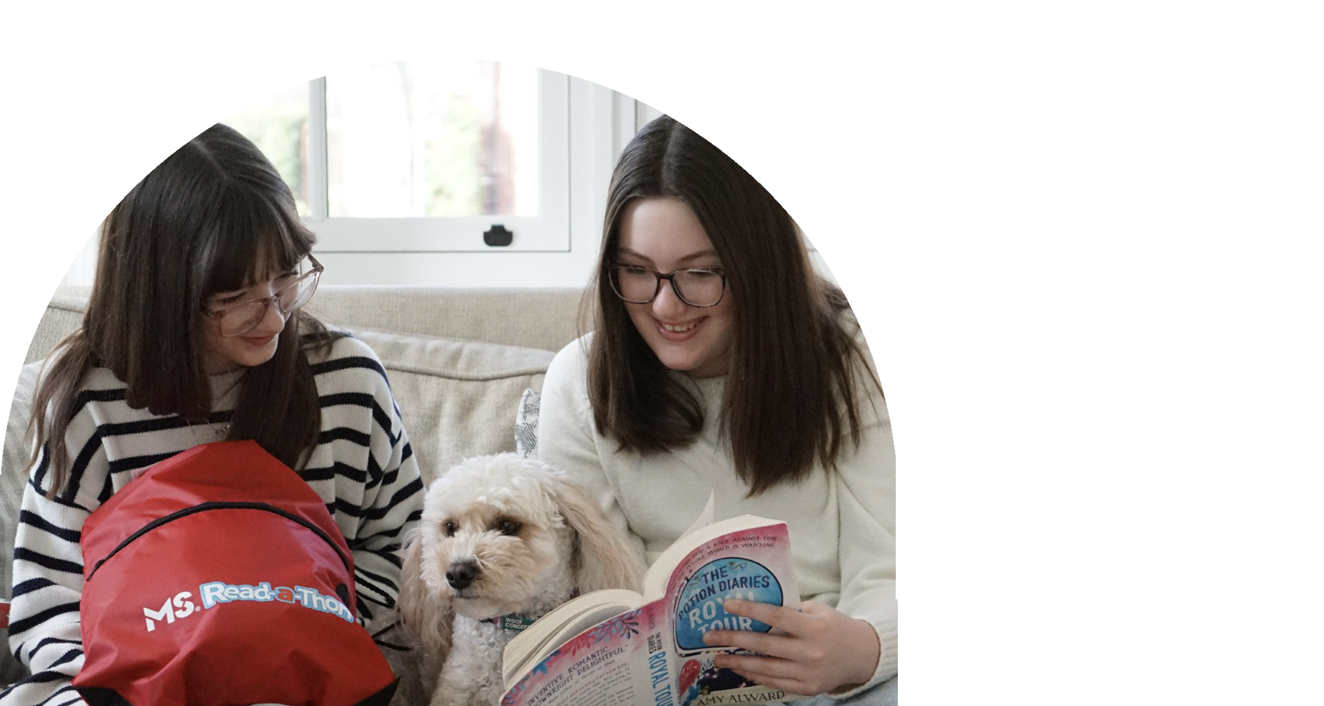 Two young girls with long brown hair hold a book out for their fuzzy white dog.