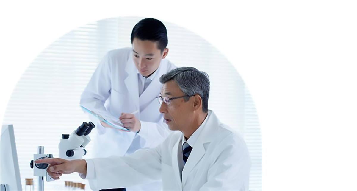 Deux chercheurs sont vêtus d’un sarrau blanc. L’un d’eux porte des lunettes et pointe du doigt un écran d’ordinateur, et l’autre tient dans ses mains une planchette à pince et un stylo. 