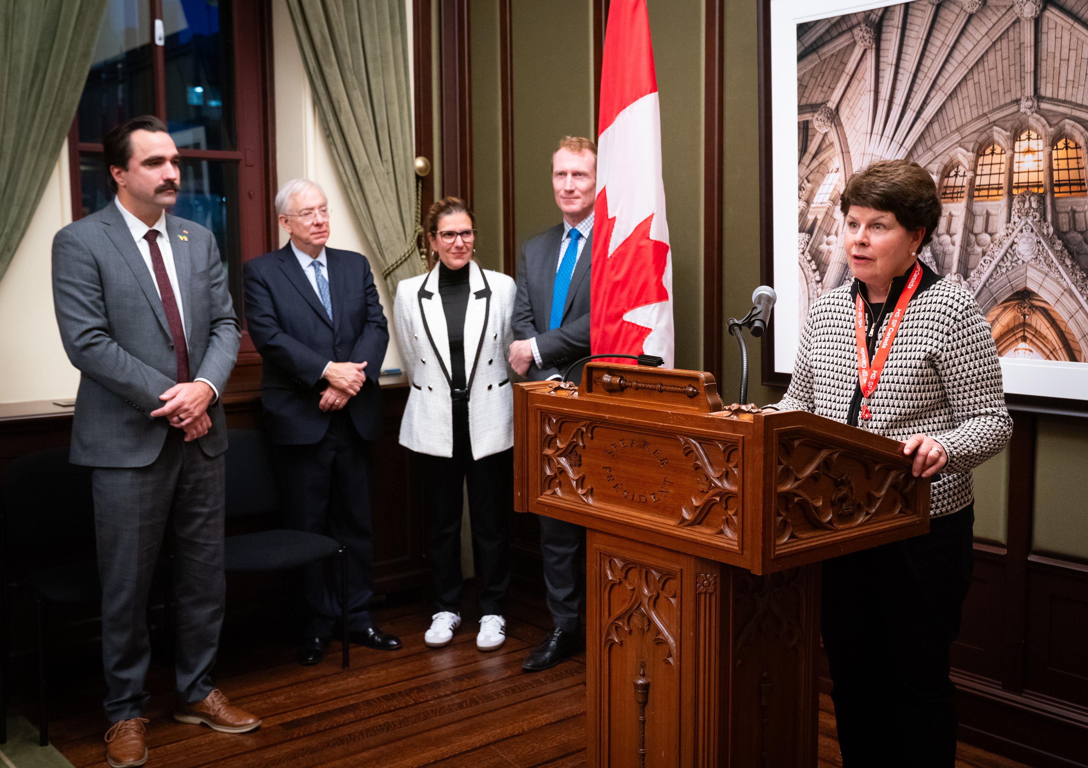 Woman speaking at a podium with a Canadian flag, while three people stand and listen nearby.