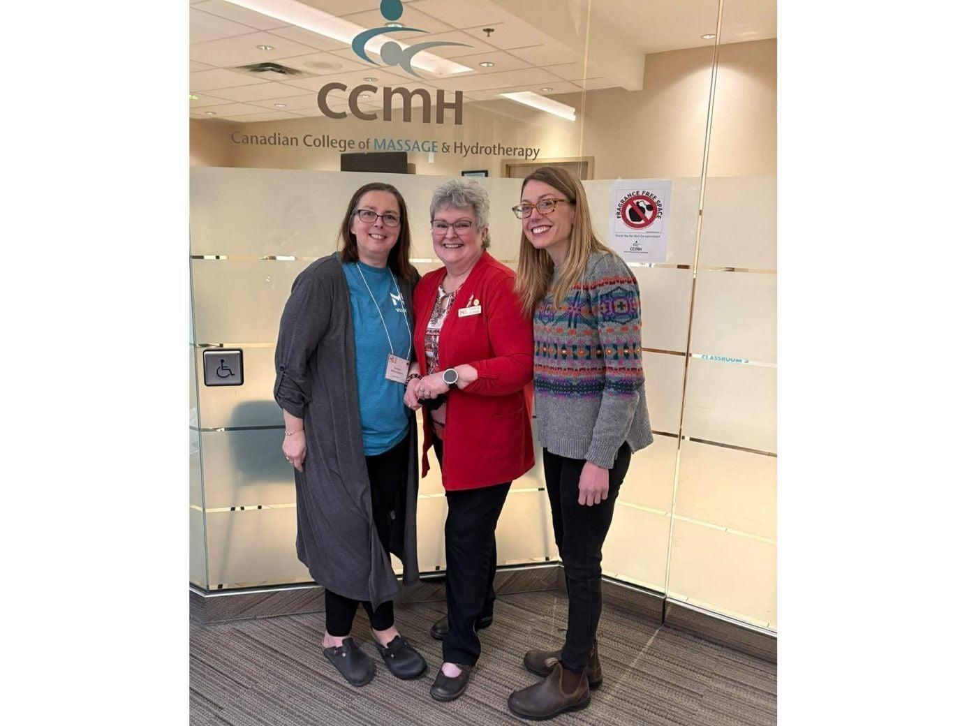 Trois femmes prennent la pose en souriant devant l’enseigne de la porte d’entrée du bureau du CCMH (Collège canadien de massage et d’hydrothérapie).