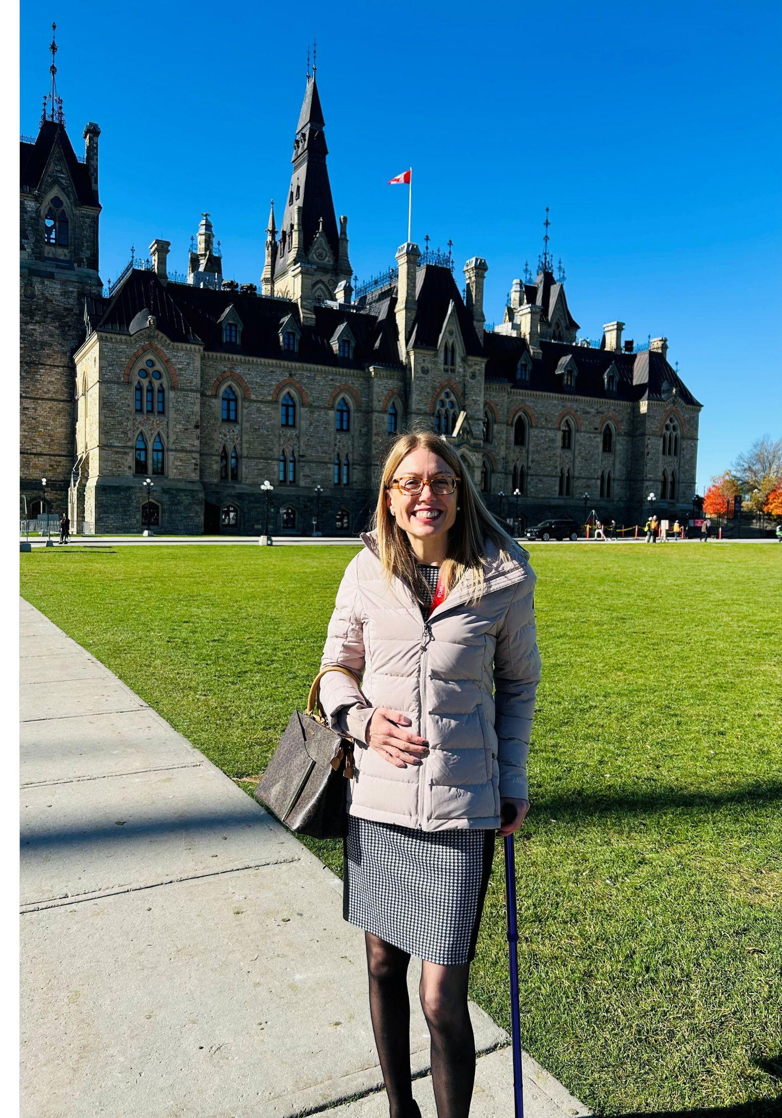 Sous un soleil radieux, Candice sourit en s’appuyant sur sa canne devant la Colline du Parlement. Elle porte des lunettes, un manteau beige et une jupe courte à carreaux.