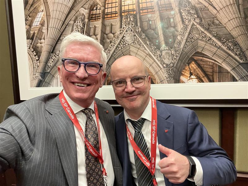 Two smiling men in suits wearing red lanyards pose together indoors.