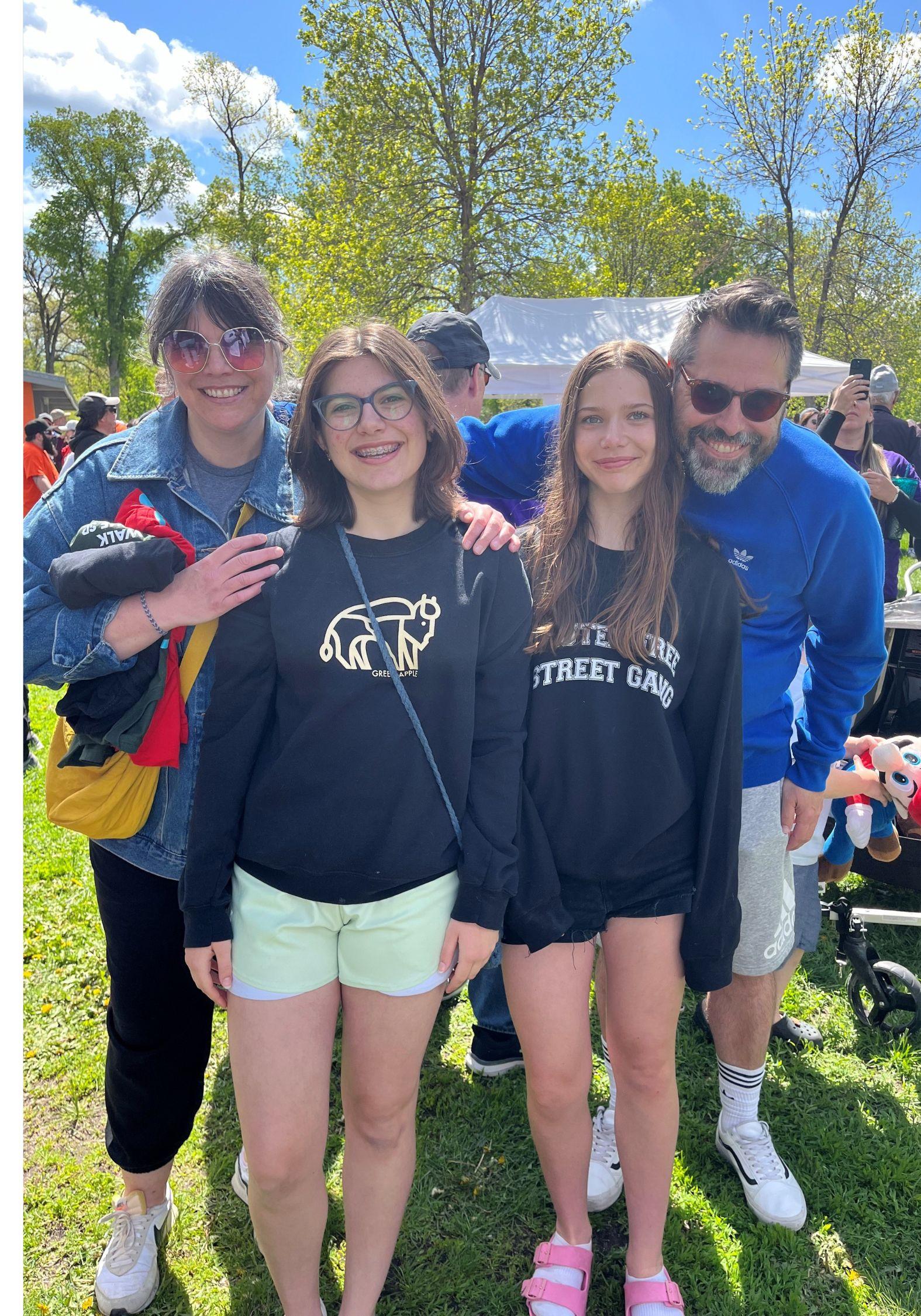 Une femme, un homme et deux jeunes filles prennent la pose avec le sourire sous un ciel ensoleillé. En arrière-plan apparaissent des arbres.