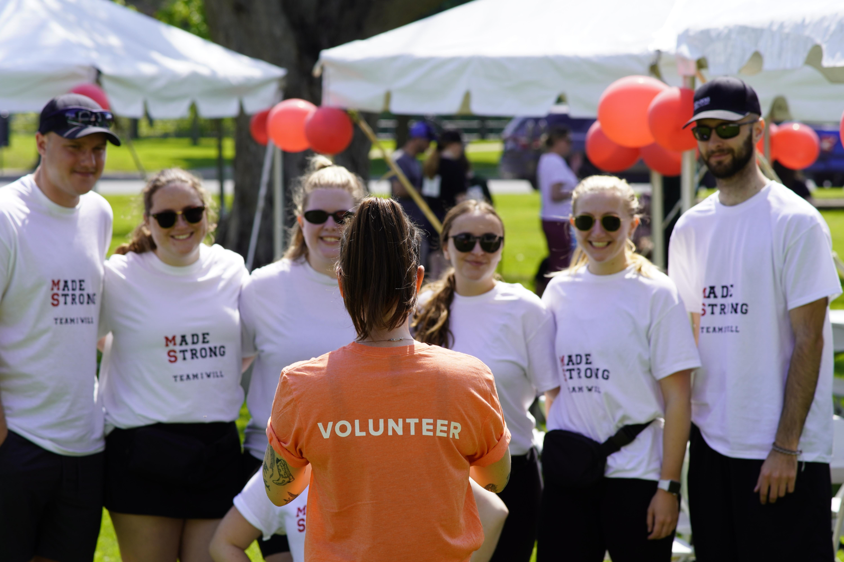 A volunteer in an orange shirt addresses a group wearing white "Make Strides" t-shirts at an outdoor event.