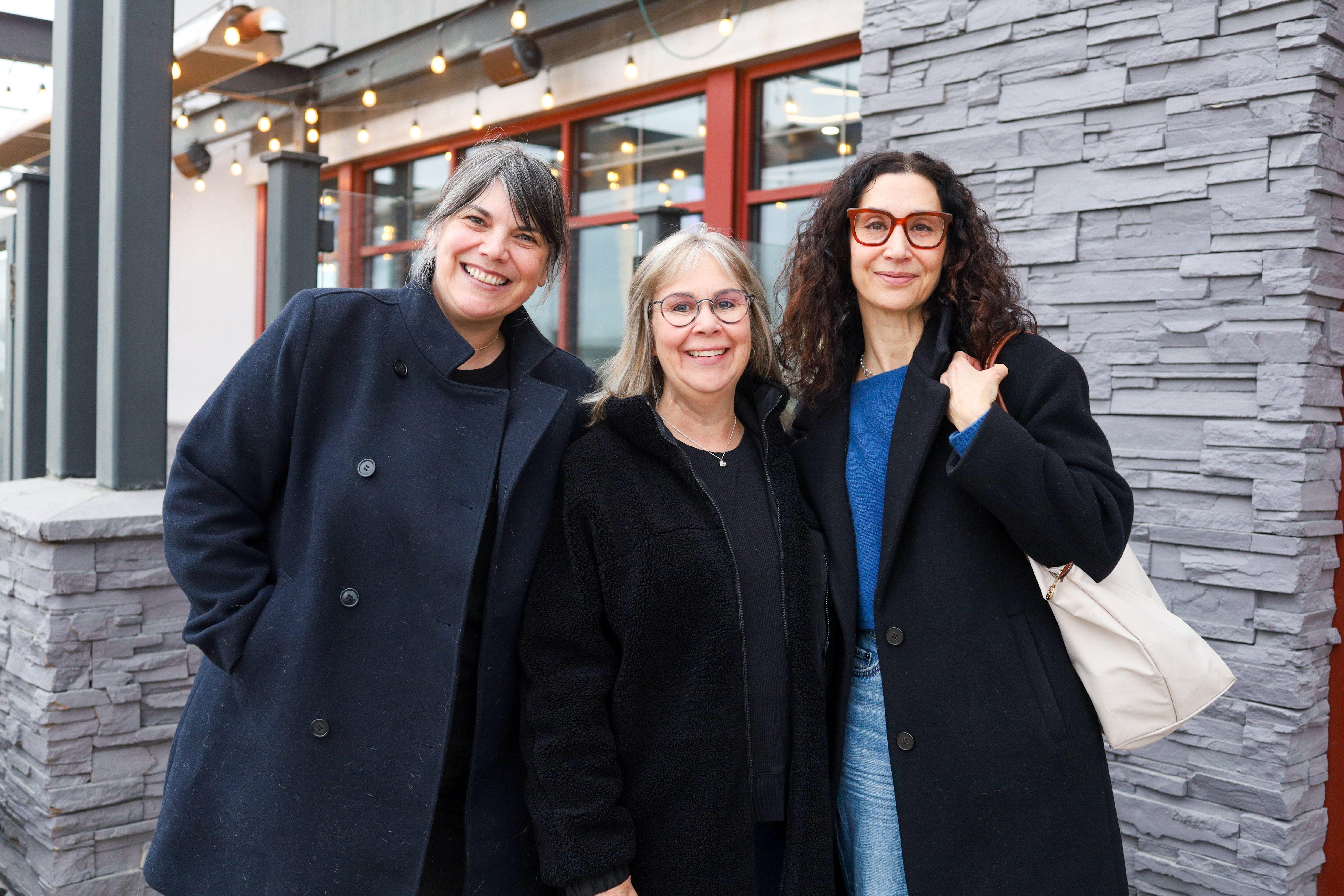 Trois femmes se tiennent debout devant un mur de pierres par un temps nuageux. Elles portent toutes un manteau de couleur foncée et elles sourient. 