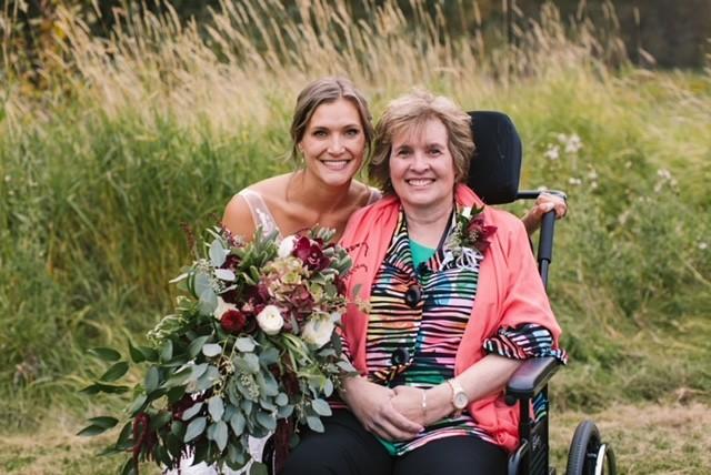 Alli tient un bouquet de fleurs. Sa mère est assise dans un fauteuil roulant. Les deux femmes sourient. 