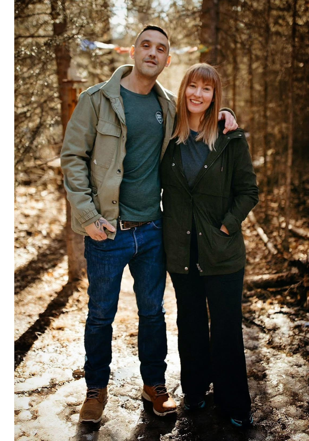 Jaclyn et son mari sourient. Ils se trouvent dans une forêt où percent les rayons du soleil, durant l’automne, et ils portent des vestes de plein air confortables.