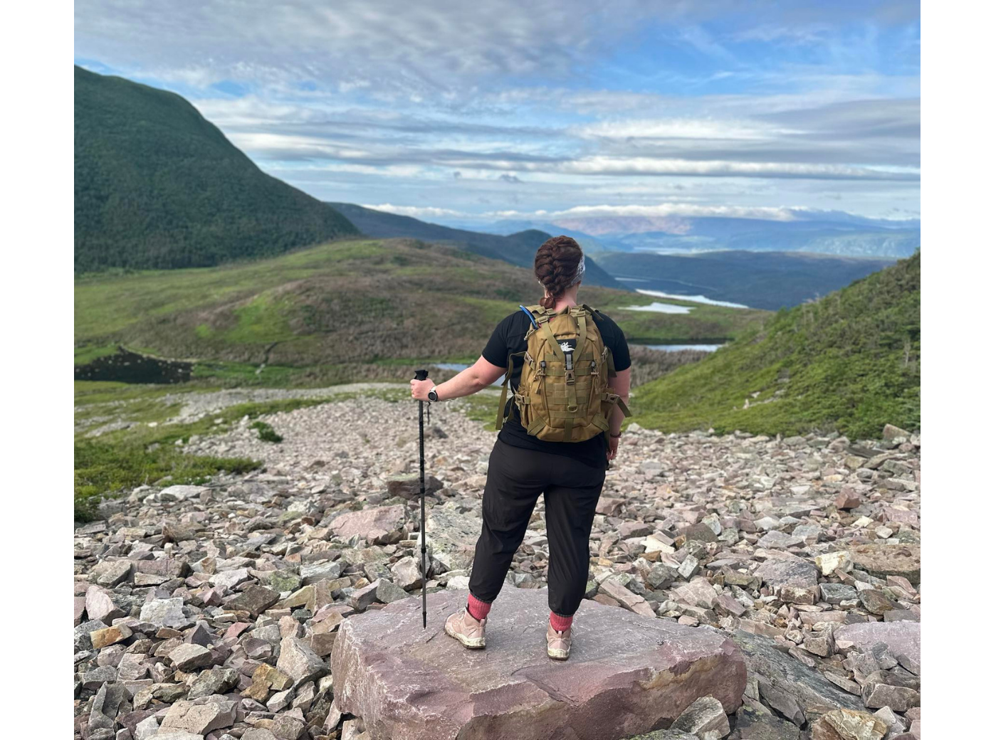 Janice, équipée d’un sac à dos jaune, se tient sur un sentier de montagne rocailleux surplombant une vallée pittoresque.