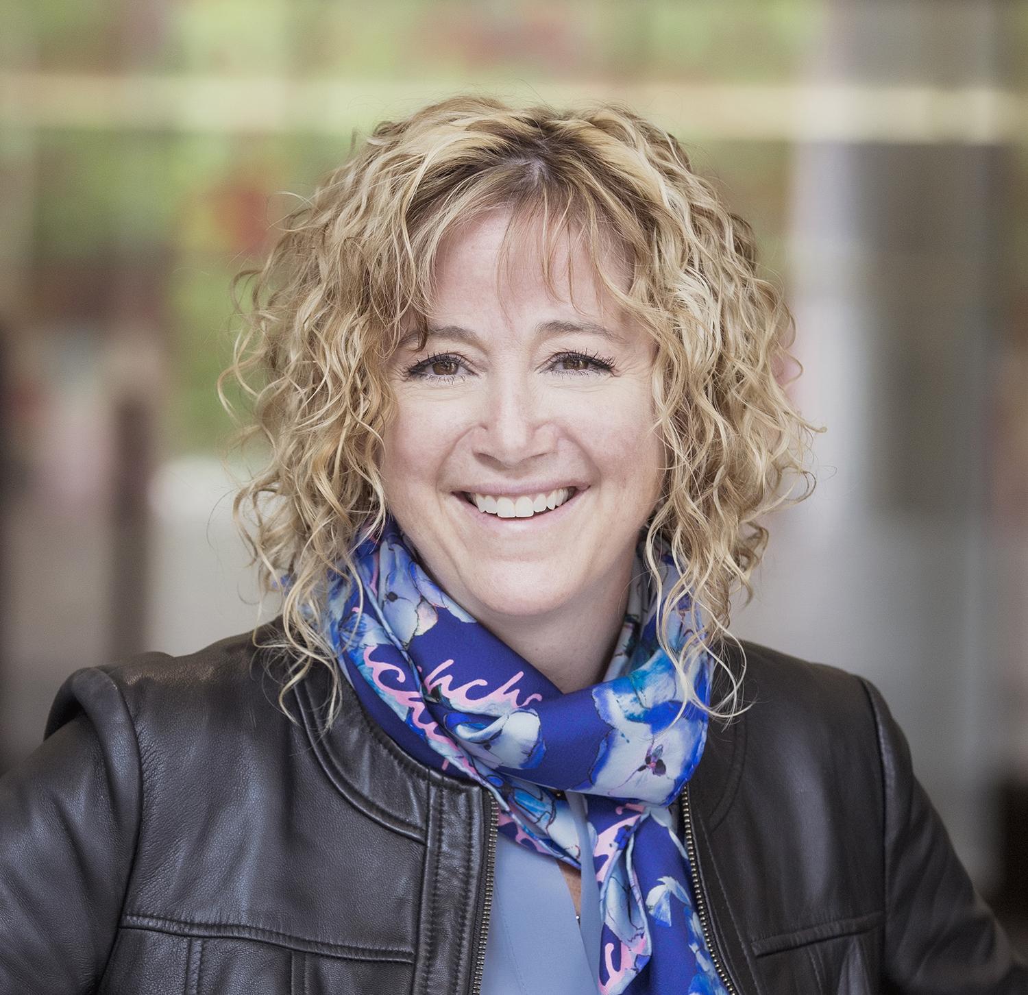 Smiling woman with curly blonde hair, wearing a leather jacket and colorful scarf.