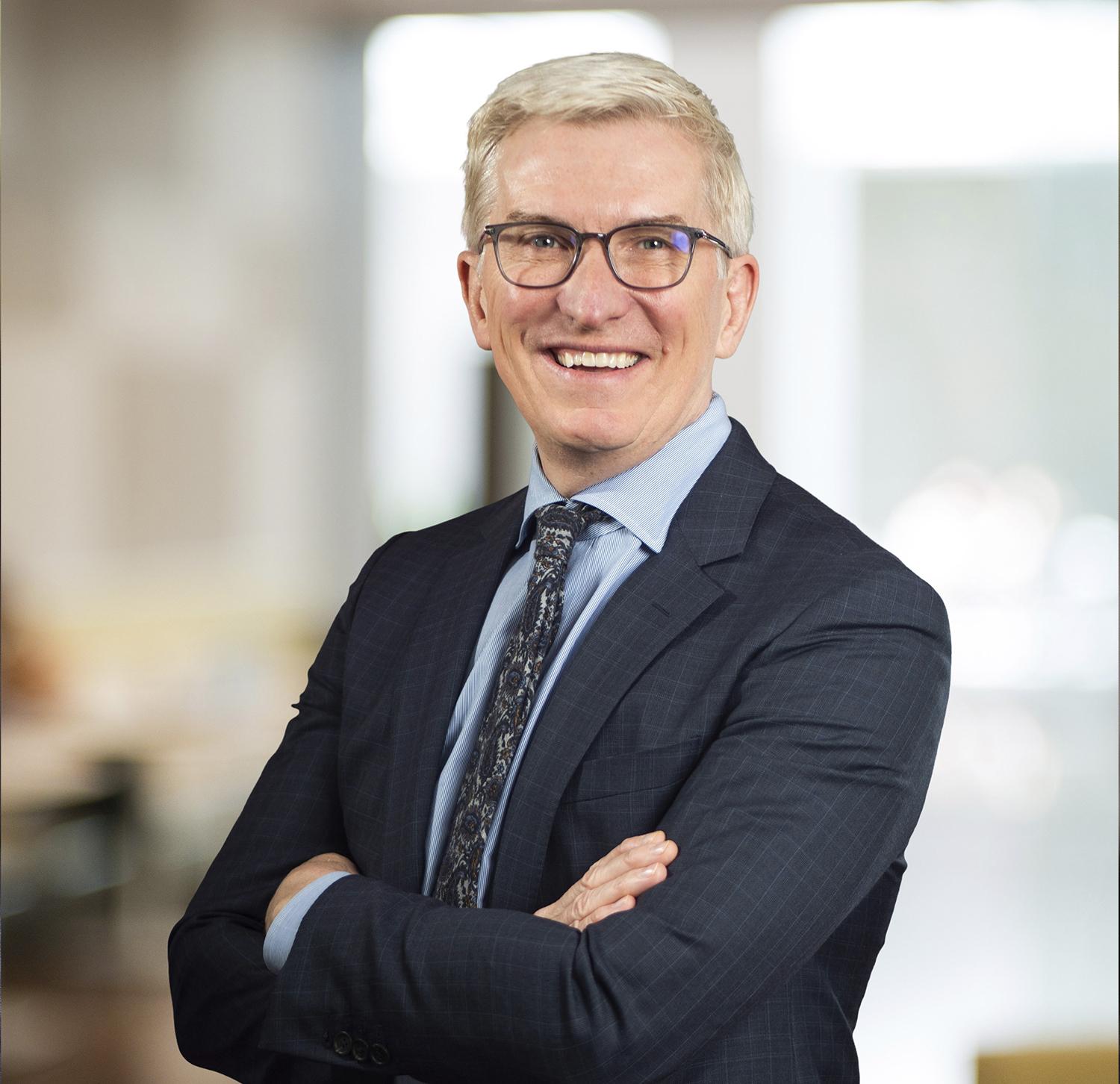 Professional headshot of a smiling middle-aged man with glasses, arms crossed, wearing a dark suit.