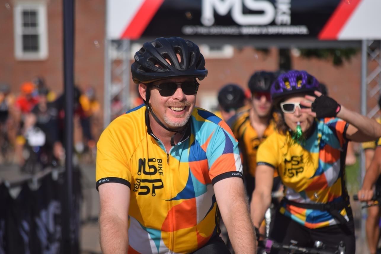 Craig est sur son vélo à la ligne de départ. Il porte un maillot du Vélo SP, un casque noir et des lunettes solaires et il sourit.