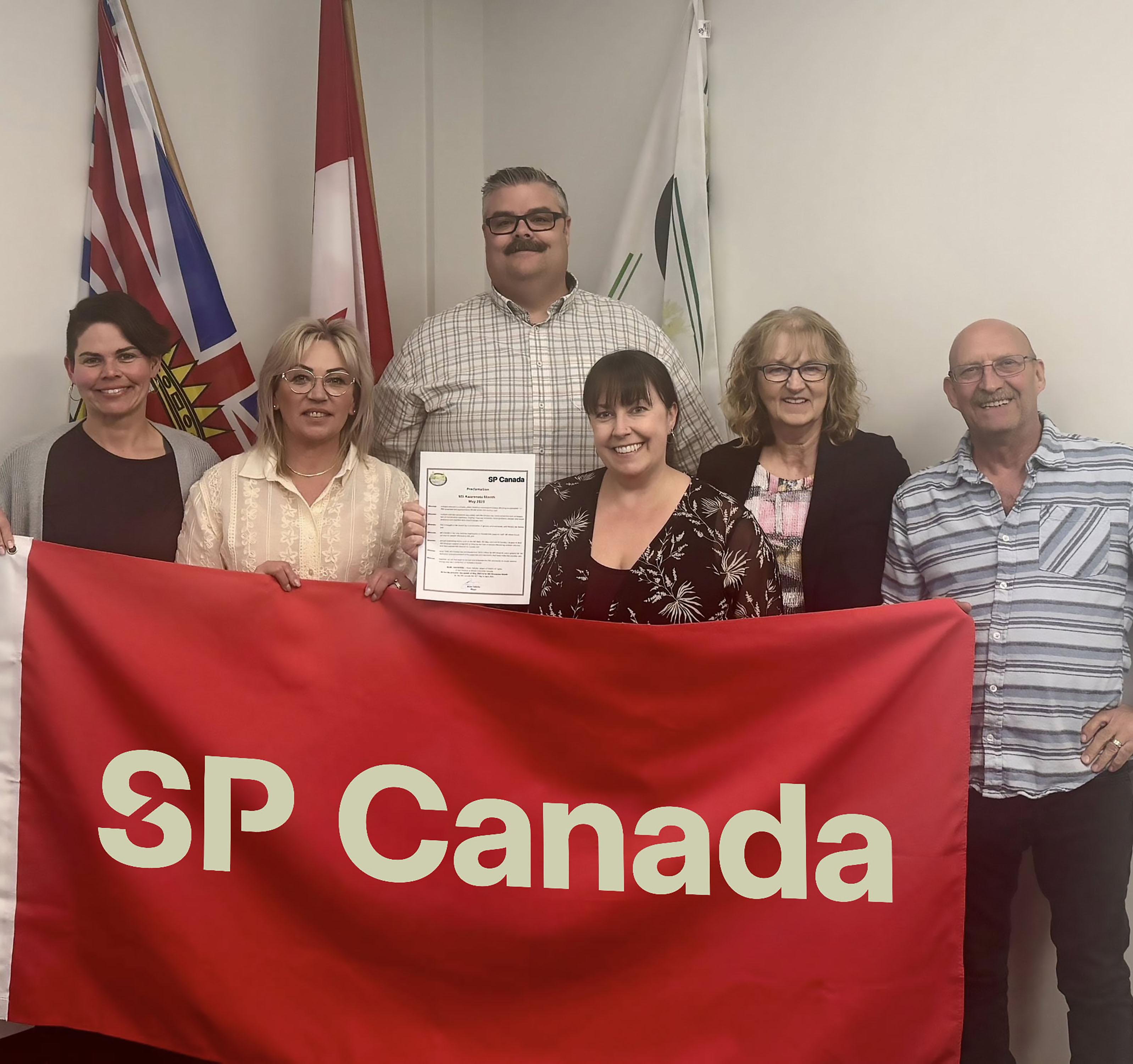 Group of people posing with a red SP Canada banner in an office setting.