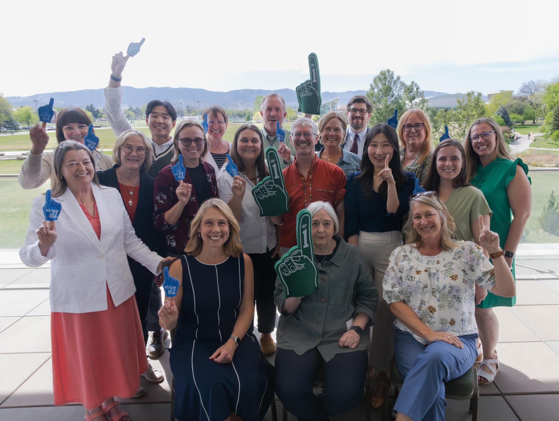 Paul et ses collègues du Département d’ergothérapie de l’Université d’État du Colorado brandissent, en signe de célébration, des mains en mousse vertes et bleues. Ce groupe pose sous un soleil radieux, devant un parc où on aperçoit des arbres.