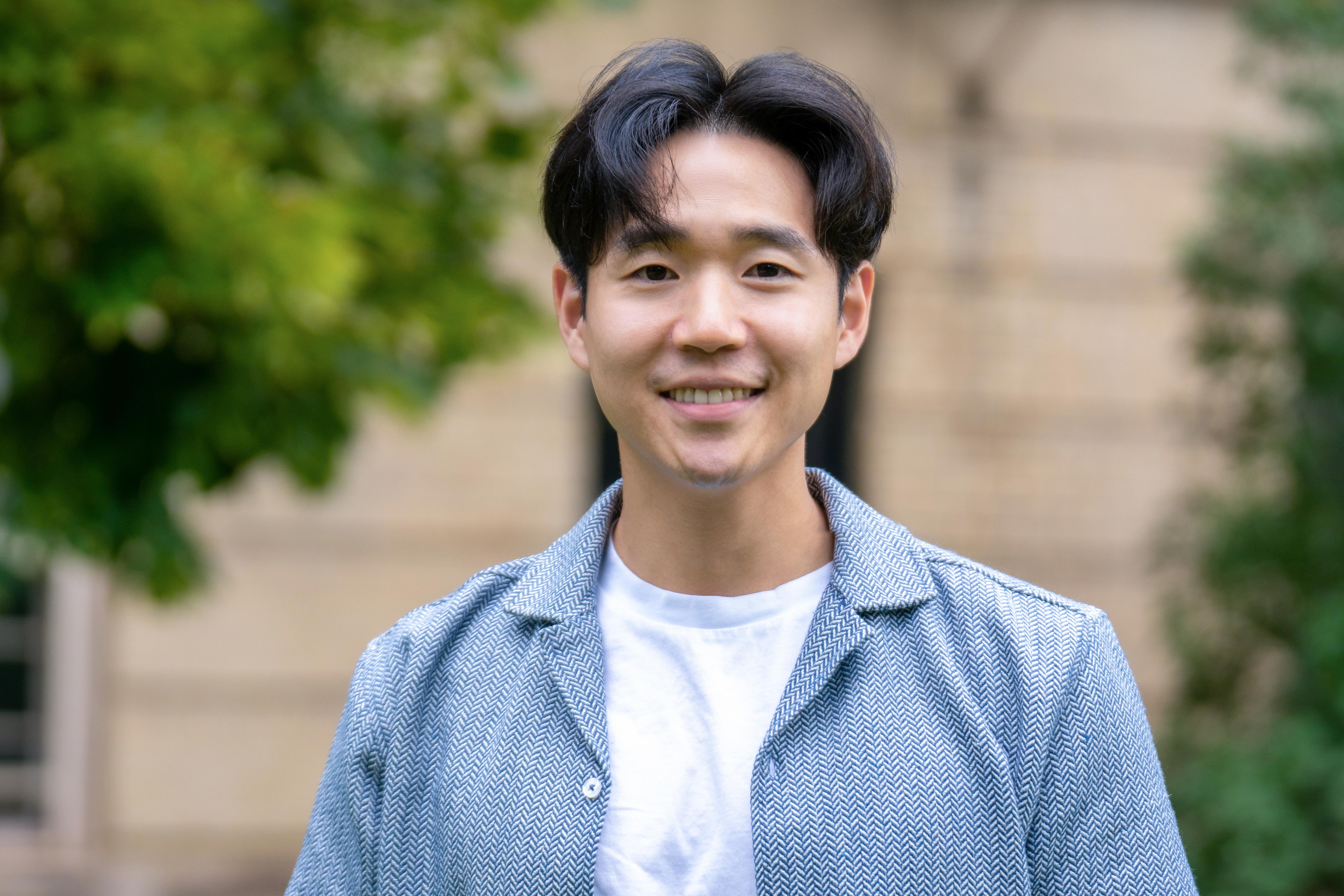 Paul Yoo, Ph. D., porte un t-shirt blanc et une veste bleu pâle. Il sourit et se tient debout devant un immeuble bordé d’arbres.