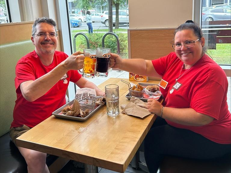 Two people in red shirts toasting drinks at a restaurant table with food.