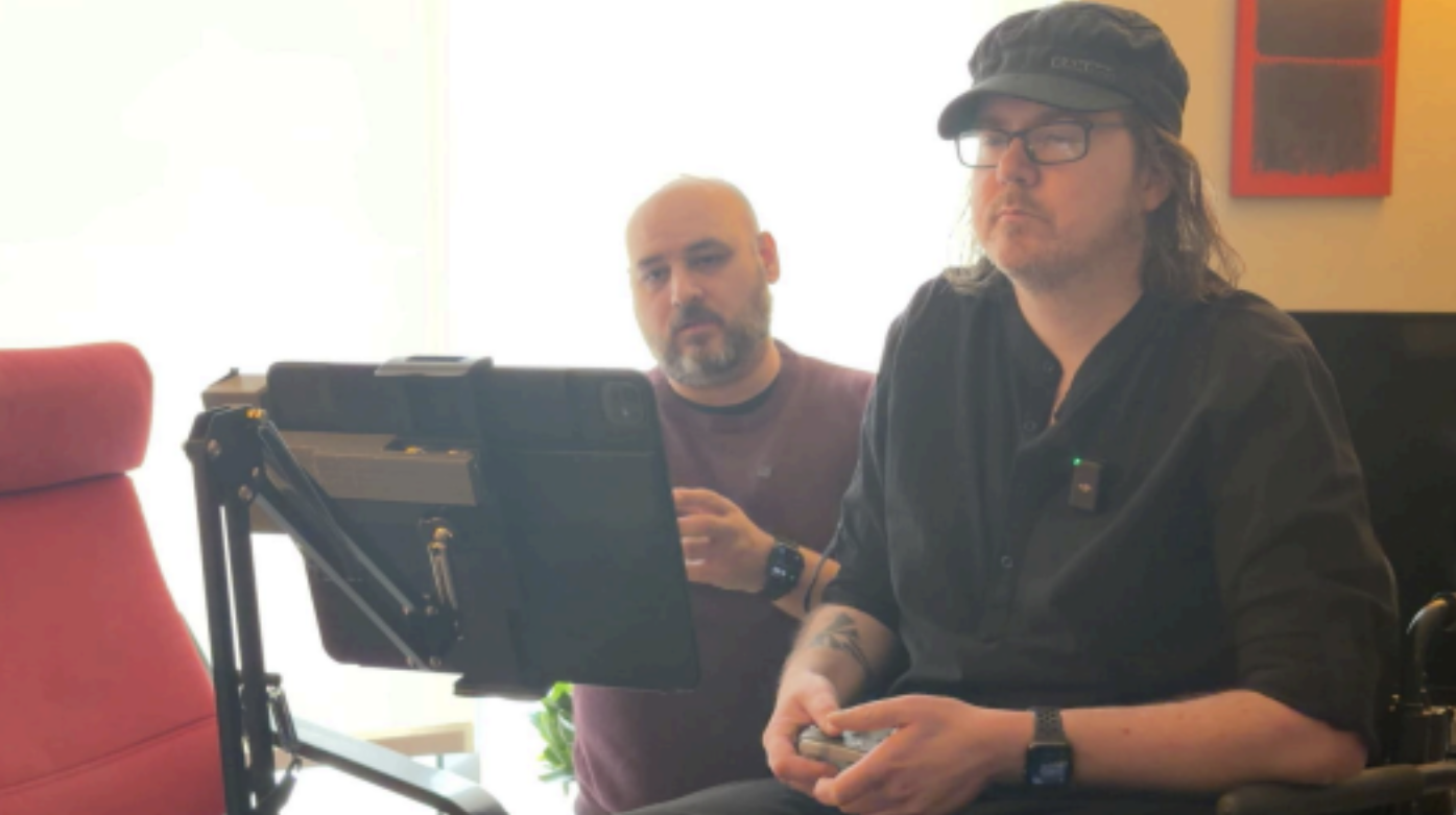 Jason et Edward sont assis devant l’écran d’une tablette adaptée, que manipule Jason à l’aide d’une manette. Jason est à l’avant-plan.