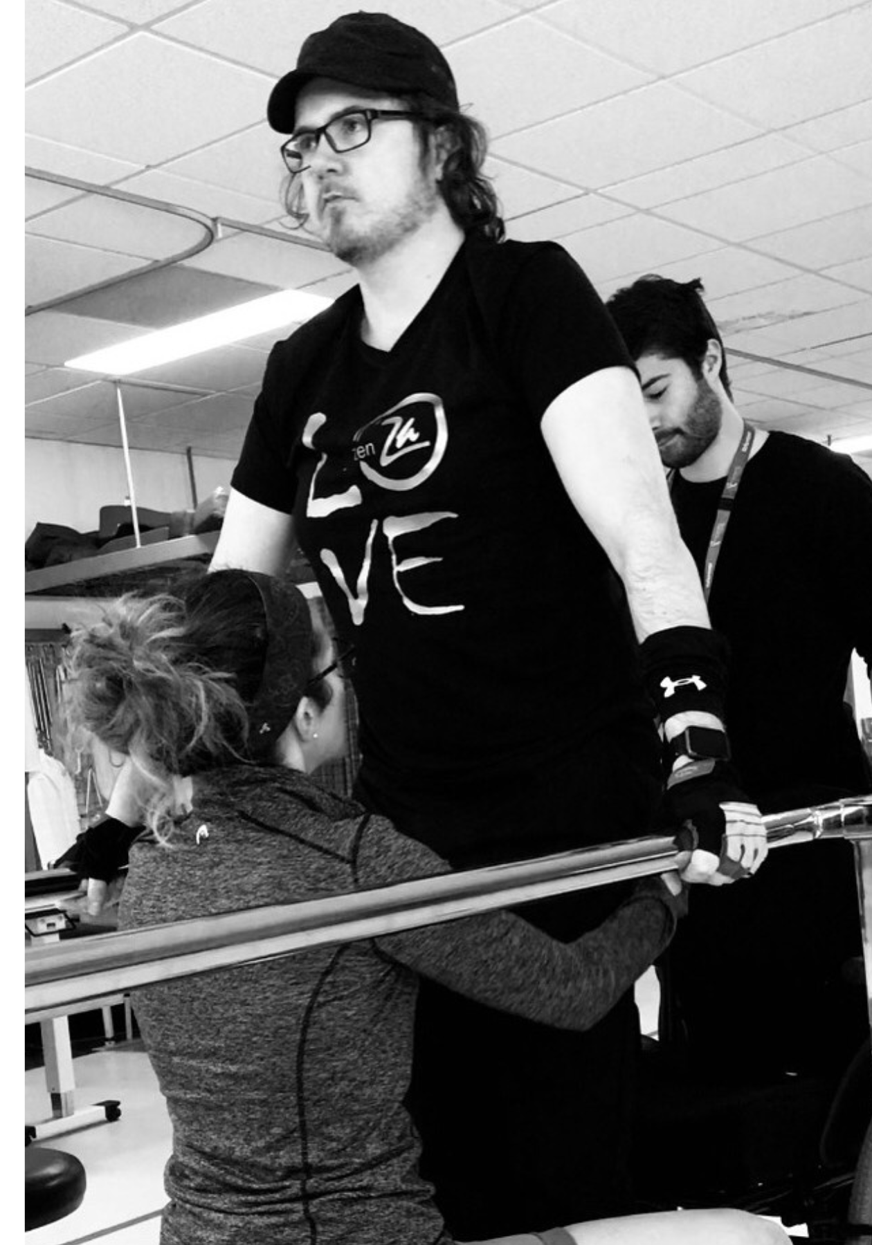 Photo en noir et blanc. Jason fait des exercices entre des barres parallèles lors d’une séance de physiothérapie. Il porte un t-shirt noir sur lequel est écrit « LOVE ». 