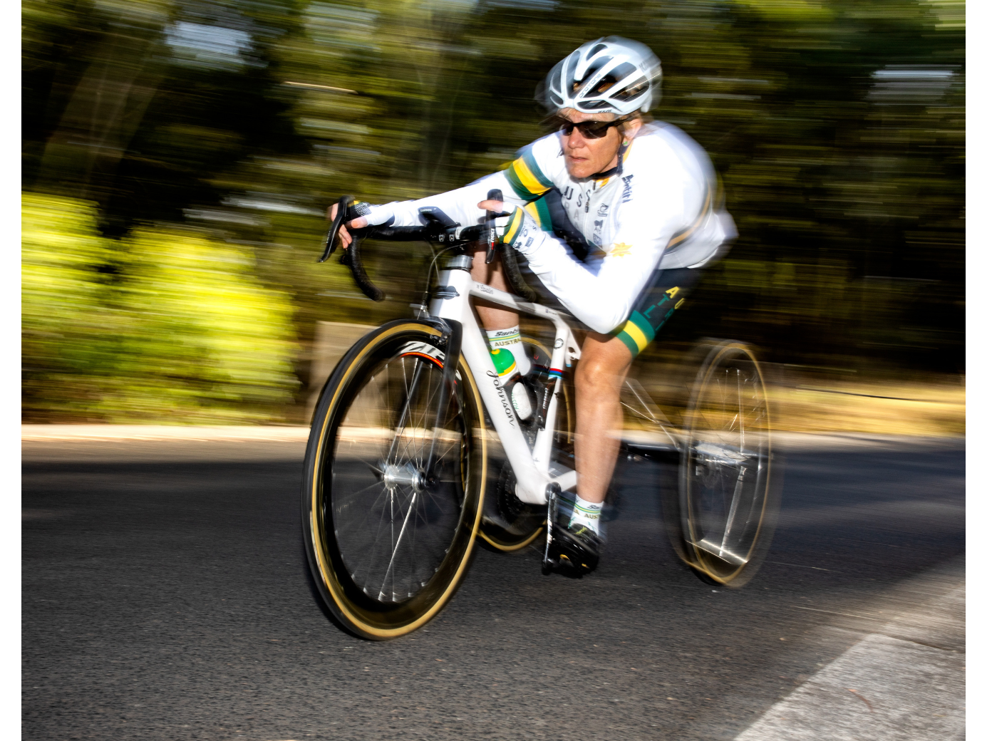 Carol porte un casque blanc et un maillot de cyclisme blanc. Elle roule à grande vitesse sur un vélo de route. L’arrière-plan est vert et flou.