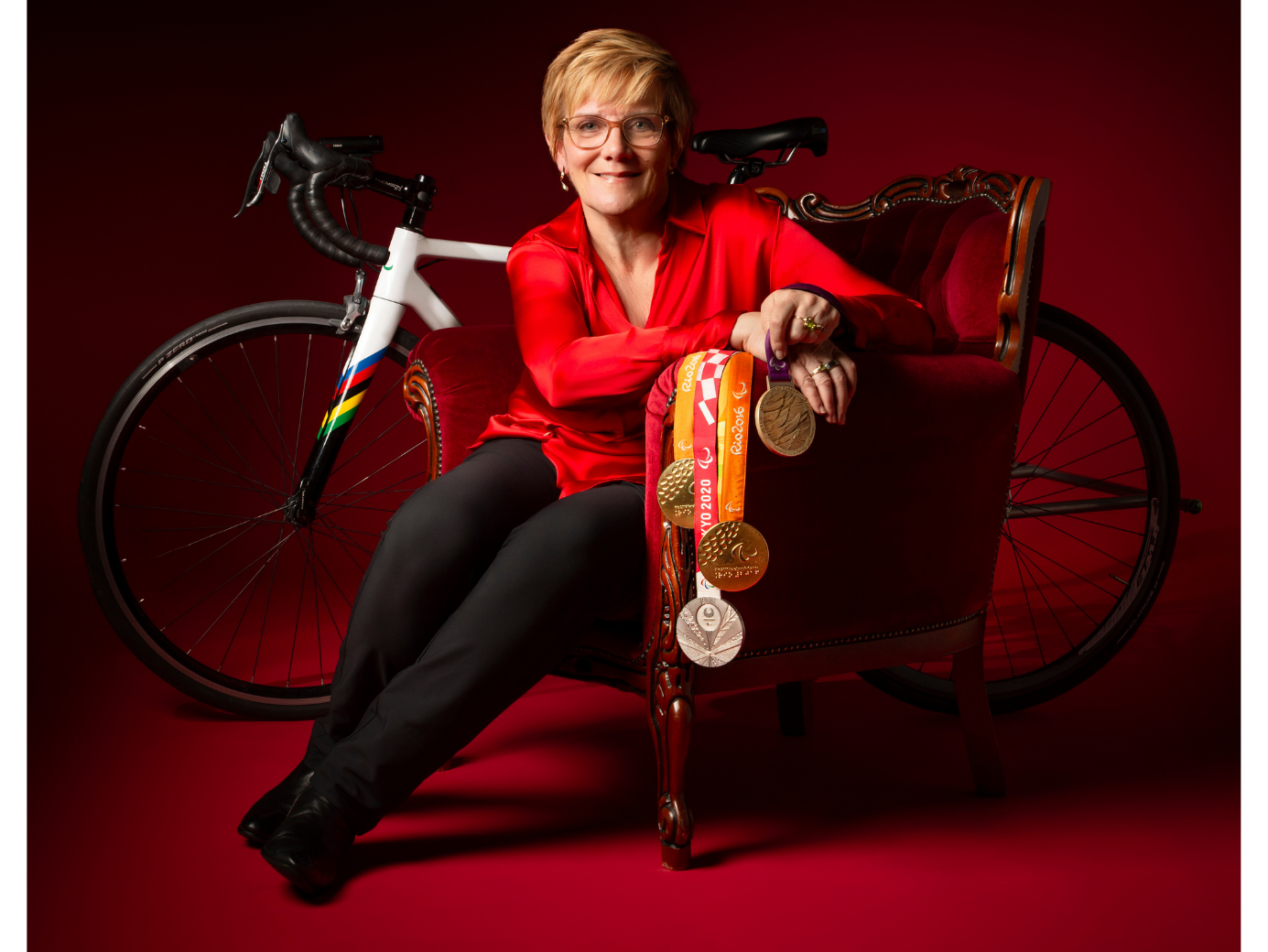 Carol porte un chemisier rouge et un pantalon noir. Elle est assise dans un fauteuil rouge devant un vélo de route et elle tient des médailles.