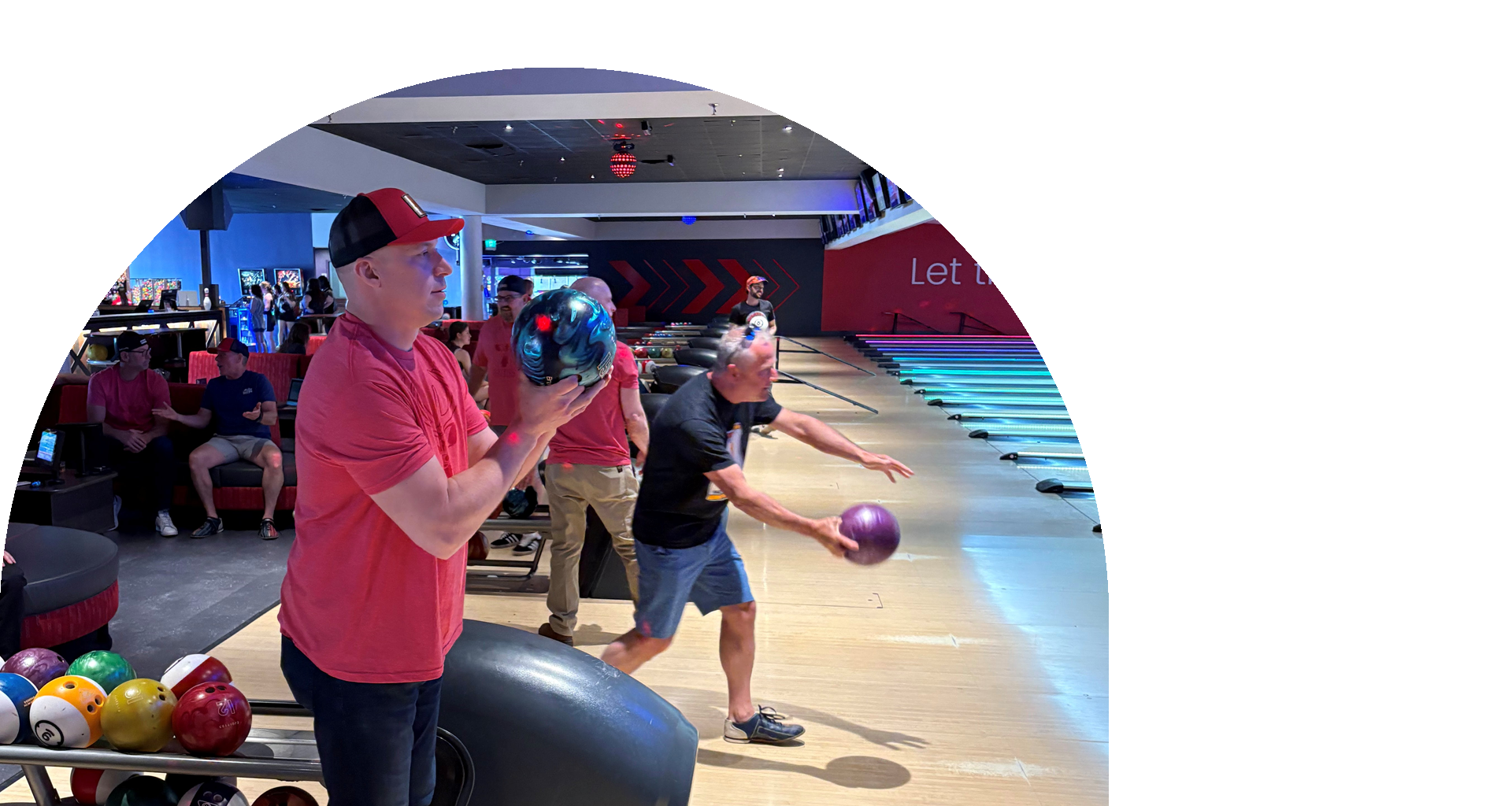 Photo of a man in a red shirt and cap holding a bowling ball in a busy bowling alley, with others bowling behind him.