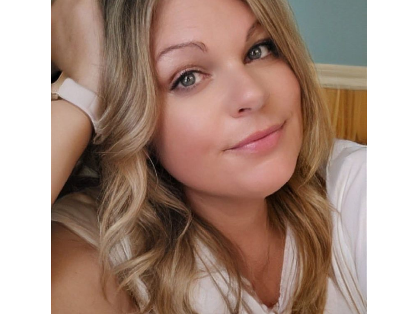 Michelle porte un t-shirt blanc. Elle a les cheveux longs, blonds et ondulés. L’un de ses bras est replié vers le haut et sa tête est appuyée sur sa main.