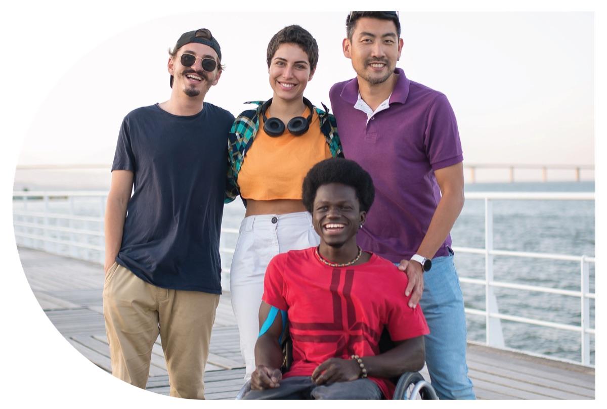 Quatre jeunes adultes souriants, dont une personne en fauteuil roulant, réunis sur une promenade au bord de l'eau.