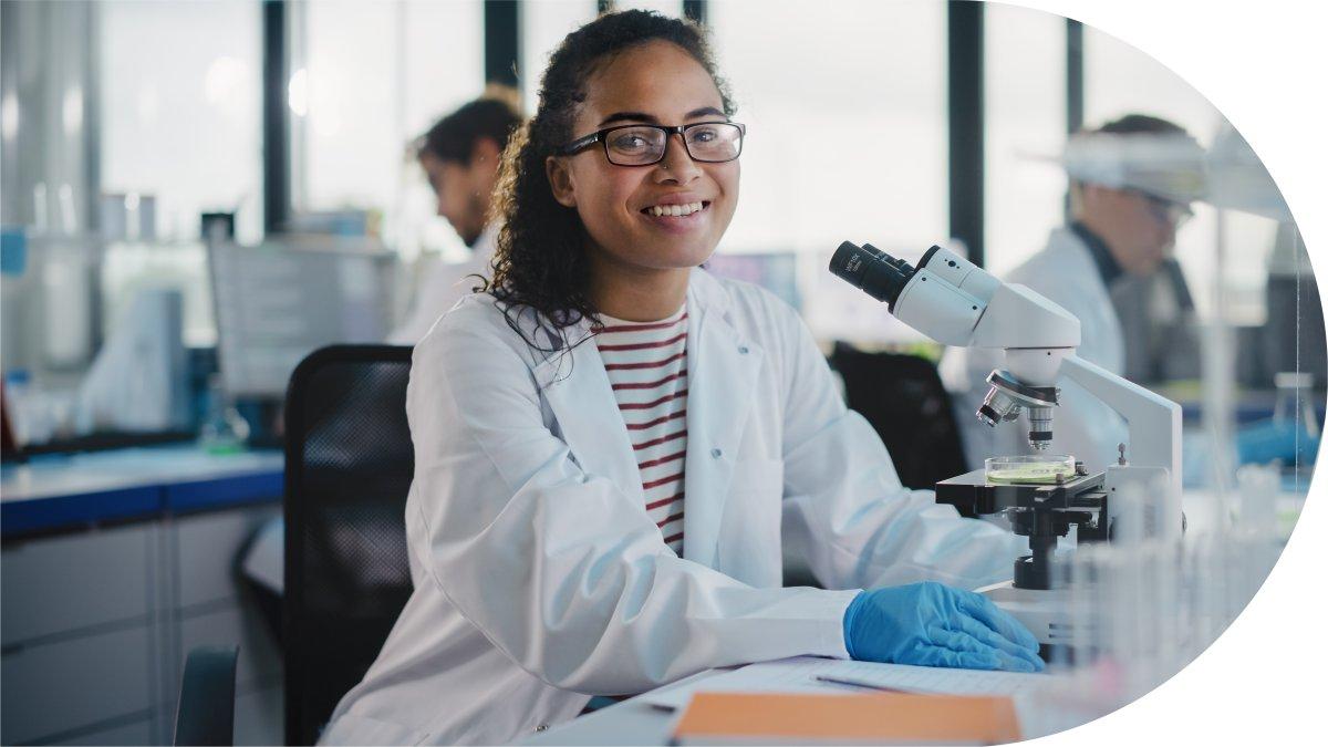 Une personne souriante en sarrau et lunettes, assise devant un microscope dans un laboratoire.