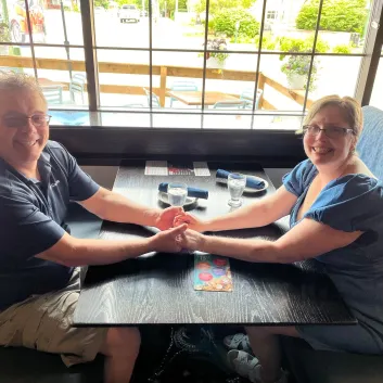 James, wearing a black shirt, and Yolanda, wearing a blue dress, sitting at a restaurant booth and holding hands.