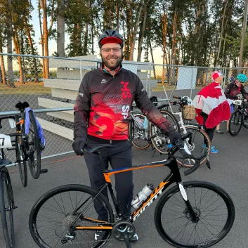 Brennan sourit et se tient debout, devant son vélo. Il porte un casque de vélo, des lunettes, des gants noirs, un pantalon noir, ainsi qu’une veste noir et rouge. En arrière-plan se trouvent d’autres vélos placés contre une clôture métallique.