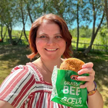 Caitlin sourit en tenant un Teen Burger™. Elle est à l’extérieur. En arrière-plan apparaissent des arbres.