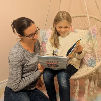 Izzy, wearing a beige sweater and jeans, sitting on a swing while holding a book beside her mother, Lindsey, wearing a grey top and jeans.