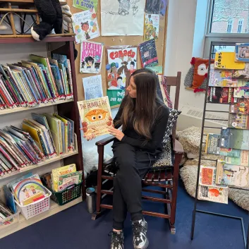 Jaclyn, wearing a black top and black pants, sitting in a chair, holding up a book.