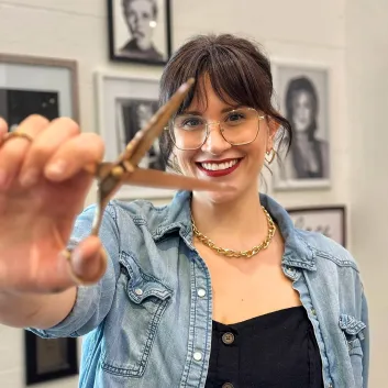 Jess, wearing a black dress and blue jacket, holding a pair of scissors.