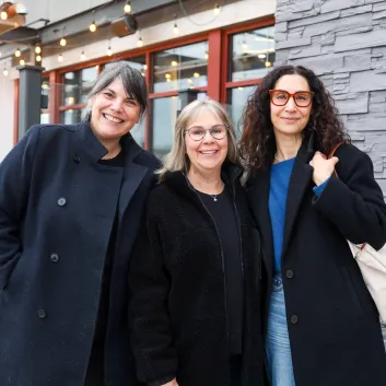 Trois femmes se tiennent debout devant un mur de pierres par un temps nuageux. Elles portent toutes un manteau de couleur foncée et elles sourient. 