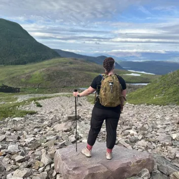Janice, équipée d’un sac à dos jaune, se tient sur un sentier de montagne rocailleux surplombant une vallée pittoresque.