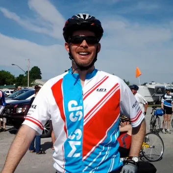 Craig porte un maillot du Vélo SP. Il est à l’extérieur et se tient devant son vélo.