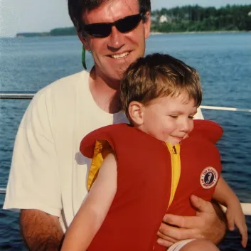 Larry porte des lunettes solaires. Assis dans un voilier, il tient dans ses bras son fils Glen, qui porte un gilet de sauvetage rouge.