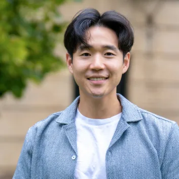 Paul Yoo, Ph. D., porte un t-shirt blanc et une veste bleu pâle. Il sourit et se tient debout devant un immeuble bordé d’arbres.