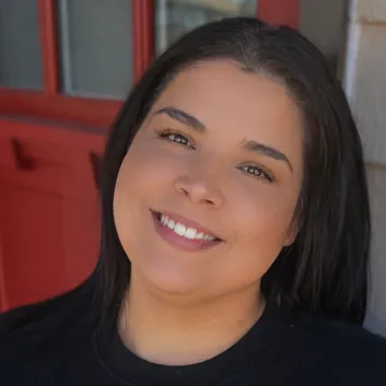 Rachel porte un chandail noir. Elle est devant une porte rouge et elle sourit.